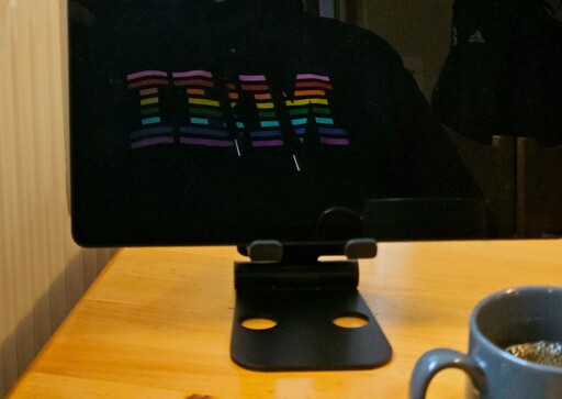 an image of a dining table, a cup of coffee, an electronic tablet in which is visible the reflection of the torso of a person wearing a hoodie, printed with the letters IBM in various colors